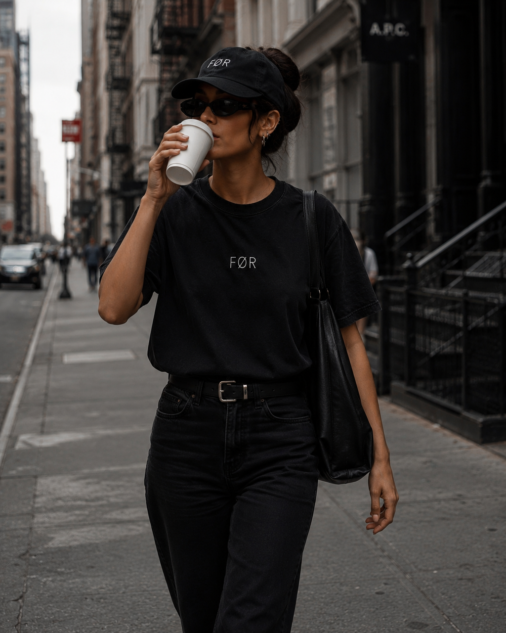 Person wearing a black outfit with 'FØR Baseball Cap and Minimal Tee, drinking from a paper cup on a city street.